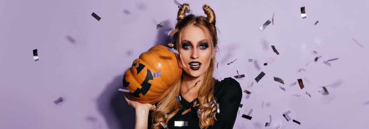 A confident young witch in a Halloween outfit posing with a pumpkin. She's also a stunning vampire girl standing against a vivid purple background